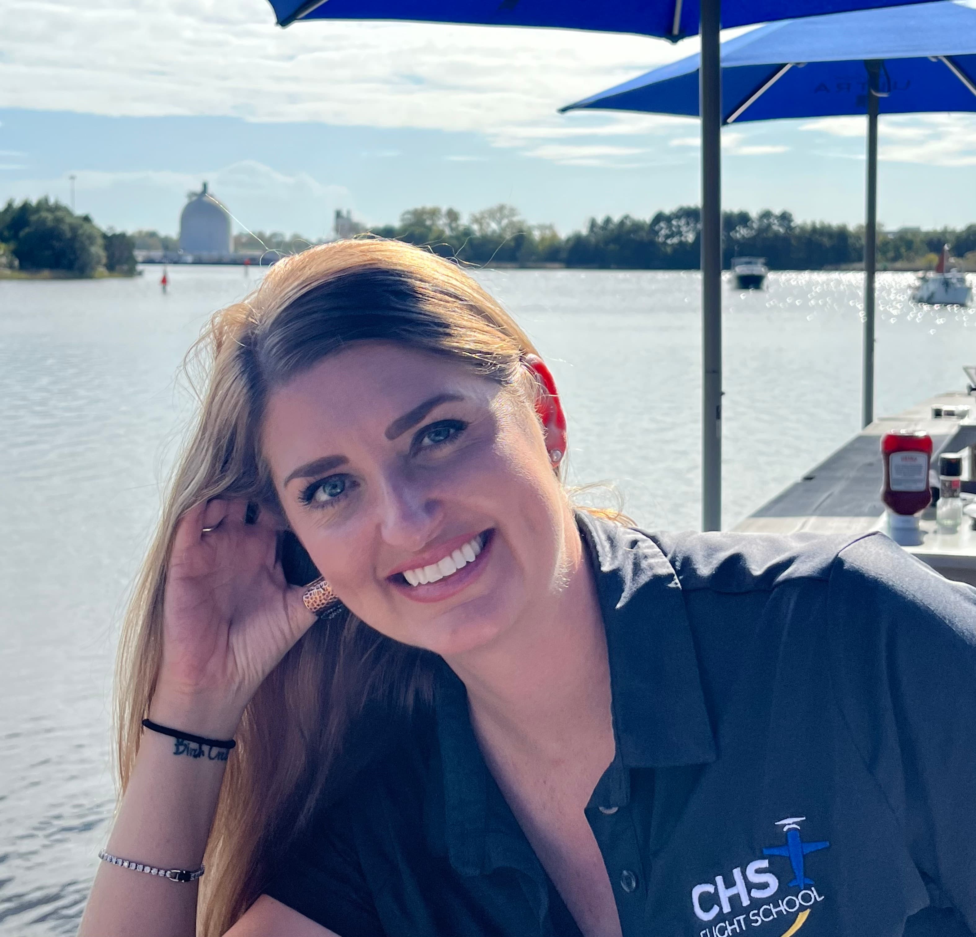 Smiling blonde woman in a CHS Flight School polo shirt sits by a sunny river.
